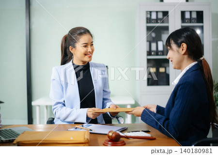 Two businesswomen in formal suits exchange cash over a contract document in an office. Bribery, fraud, and legal corruption concept with law props like gavel and folders. Two businesswomen in formal suits exchange cash over a contract document in an office. Bribery, fraud, and legal corruption concept with law props like gavel and folders. 140180981