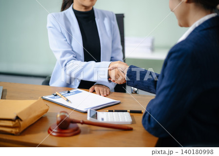 Two professional women shake hands over a signed contract at a legal office desk, symbolizing trust, agreement, and partnership. Two professional women shake hands over a signed contract at a legal office desk, symbolizing trust, agreement, and partnership. 140180998