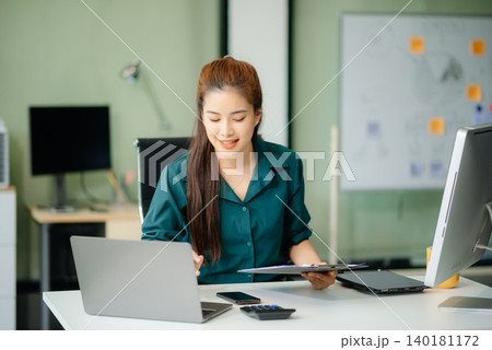 Asian woman, smiling and working at her desk in an office environment. She is wearing a business suit 140181172