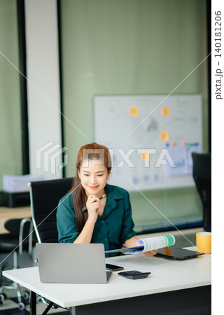 Asian woman, smiling and working at her desk in an office environment. She is wearing a business suit Asian woman, smiling and working at her desk in an office environment. She is wearing a business suit 140181206