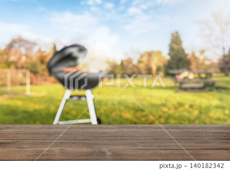 Wooden table with blurred barbecue grill in backyard garden Wooden table with blurred barbecue grill in backyard garden 140182342