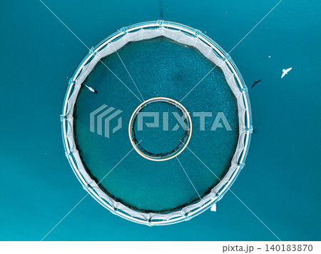 Aerial view of a fish farm located in a calm coastal bay, with floating cages arranged on the water surface. Aerial view of a fish farm located in a calm coastal bay, with floating cages arranged on the water surface. 140183870