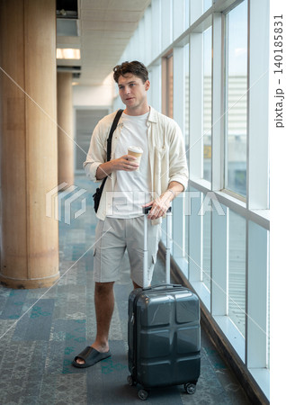 White caucasian man tourist holding coffee cup dragging luggage while standing or waiting in airport White caucasian man tourist holding coffee cup dragging luggage while standing or waiting in airport 140185831