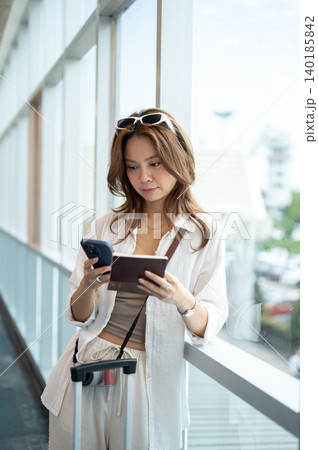 Pretty asian woman looking at phone holding passport as standing at glass wall in airport's building 140185842