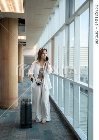 Pretty asian woman tourist talking on phone dragging a luggage while walking in airport's building. Pretty asian woman tourist talking on phone dragging a luggage while walking in airport's building. 140185851