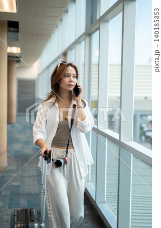 Pretty asian woman tourist talking on phone as dragging luggage while walking in airport's building. Pretty asian woman tourist talking on phone as dragging luggage while walking in airport's building. 140185853