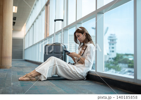 Bored asian woman holding passport looking at phone sitting on floor waiting in airport's building. 140185858