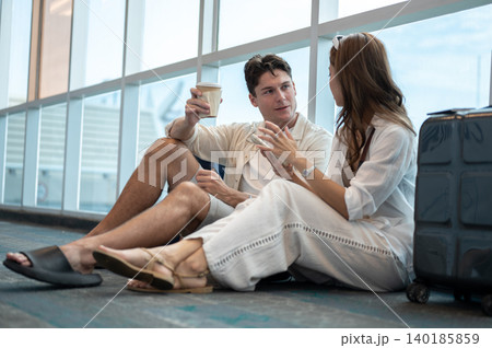 Asian woman girlfriend talking to white man boyfriend holding coffee sitting on floor in the airport 140185859
