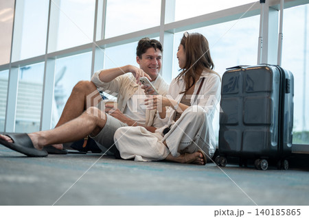 Asian woman girlfriend talking to white man boyfriend while sitting with luggage on floor in airport 140185865