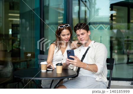 White man boyfriend holding phone showing to asian woman girlfriend sitting at table in coffee shop. 140185999