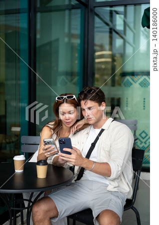 Asian woman girlfriend and white man boyfriend looking at phone sitting at table in coffee shop cafe 140186000