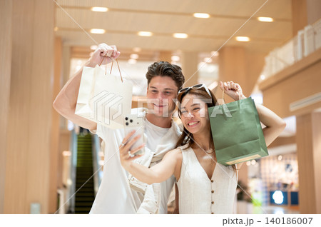 Asian woman girlfriend taking photo on phone with white man boyfriend holding shopping bag in mall. Asian woman girlfriend taking photo on phone with white man boyfriend holding shopping bag in mall. 140186007