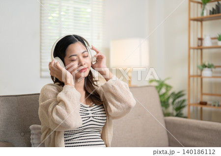 Pregnant asian woman mother closing eyes listening to music as sitting on living room's sofa at home Pregnant asian woman mother closing eyes listening to music as sitting on living room's sofa at home 140186112