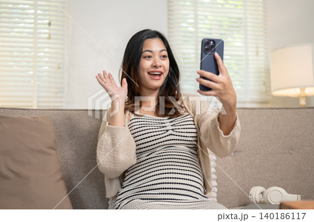 Surprised pregnant asian woman looking at phone on video call sitting on living room's sofa at home. Surprised pregnant asian woman looking at phone on video call sitting on living room's sofa at home. 140186117