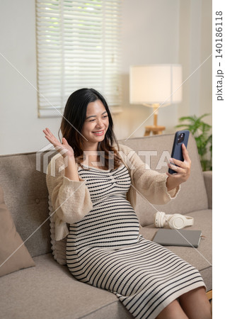 Happy pregnant asian woman looking at phone on video call while sitting on sofa at home living room. 140186118
