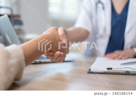 Close up of woman patient shaking hands with a doctor in meeting at wooden table in hospital clinic. Close up of woman patient shaking hands with a doctor in meeting at wooden table in hospital clinic. 140186154