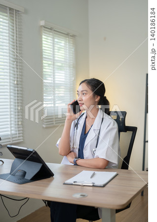Young asian woman doctor folding arm talking on phone sitting at working table in hospital or clinic Young asian woman doctor folding arm talking on phone sitting at working table in hospital or clinic 140186184