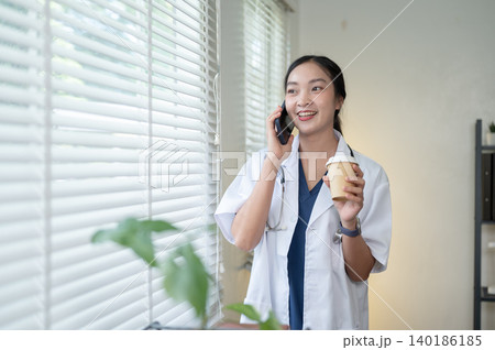 Pretty asian woman doctor holding coffee talking on phone standing beside window in hospital clinic. 140186185