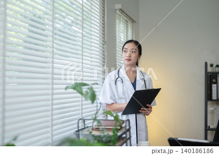 Pretty asian woman doctor holding clipboard standing looking out of a window in hospital or clinic. Pretty asian woman doctor holding clipboard standing looking out of a window in hospital or clinic. 140186186