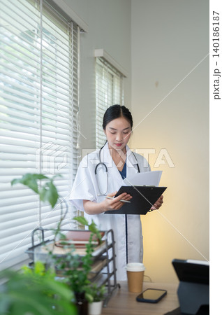 Pretty asian woman doctor reading papers on clipboard standing beside window in hospital or clinic. 140186187
