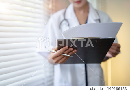 Close up of young woman doctor reading papers on clipboard standing beside window in hospital clinic 140186188