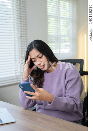 Troubled asian woman looking at phone beside laptop while sitting at wooden table in home office. Troubled asian woman looking at phone beside laptop while sitting at wooden table in home office. 140186259