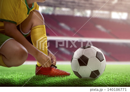 A young football player in a yellow uniform ties his red cleats on the green grass pitch next to a soccer ball in a stadium A young football player in a yellow uniform ties his red cleats on the green grass pitch next to a soccer ball in a stadium 140186771