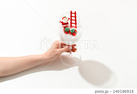 Female hand holding a clear wine glass with a miniature Santa Claus climbing a small ladder and gift boxes, against a clean white background for Christmas lifestyle advertising. 140186918