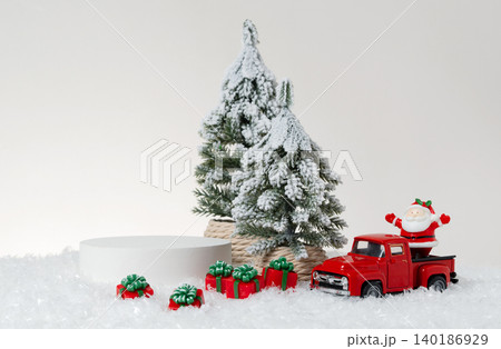 Festive Christmas product display photography featuring a white cylindrical podium, snow-covered pine tree, red truck, Santa Claus figurine, and wrapped gift boxes arranged on artificial snow. Festive Christmas product display photography featuring a white cylindrical podium, snow-covered pine tree, red truck, Santa Claus figurine, and wrapped gift boxes arranged on artificial snow. 140186929