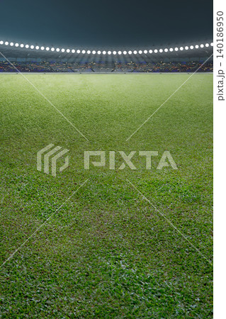 A vast, empty green grass field inside a professional sports stadium at night. Bright floodlights illuminate the pitch, with a blurred audience in the background stands A vast, empty green grass field inside a professional sports stadium at night. Bright floodlights illuminate the pitch, with a blurred audience in the background stands 140186950