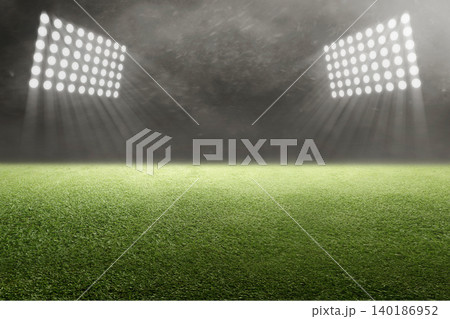 A wide-angle view of an empty green sports field at night, illuminated by powerful stadium floodlights through a hazy, dramatic atmosphere 140186952