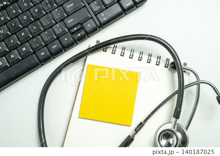 A top-down view of a medical stethoscope lying on a spiral notebook with a yellow sticky note next to a black computer keyboard, representing modern healthcare and administration 140187025