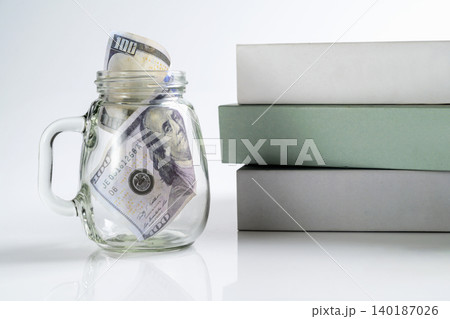 US dollar bills in a glass jar next to a stack of books, representing savings for education, student loans, tuition fees, and financial planning for university US dollar bills in a glass jar next to a stack of books, representing savings for education, student loans, tuition fees, and financial planning for university 140187026