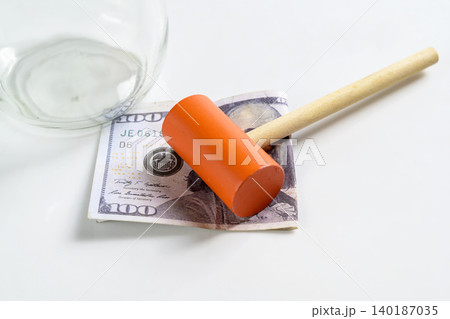 A small toy hammer lies on a hundred-dollar bill next to an empty glass jar, representing themes of broken savings, financial crisis, or emergency spending A small toy hammer lies on a hundred-dollar bill next to an empty glass jar, representing themes of broken savings, financial crisis, or emergency spending 140187035