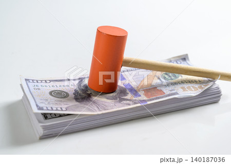 A wooden toy gavel mallet with an orange head rests on top of a stack of hundred US dollar bills, symbolizing legal costs, court fees, judgment, and auctions 140187036