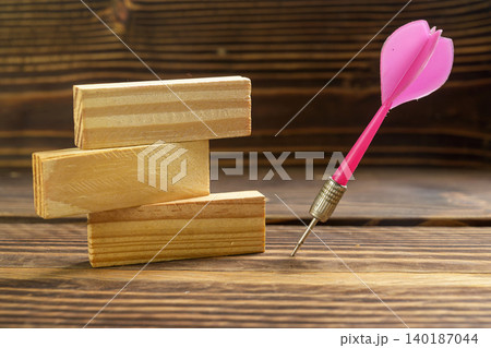 A stack of three wooden blocks next to a bright pink dart stuck into a rustic wooden surface, representing business goals, strategy, targeting, and step-by-step progress A stack of three wooden blocks next to a bright pink dart stuck into a rustic wooden surface, representing business goals, strategy, targeting, and step-by-step progress 140187044