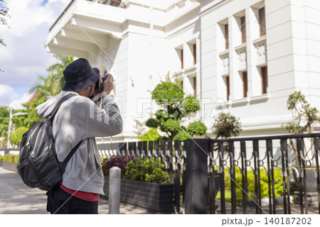 Side view of a young Indonesian southeast asian male photographer with a backpack taking a photo of a white colonial-style building on a sunny day. Travel or vacation concept Side view of a young Indonesian southeast asian male photographer with a backpack taking a photo of a white colonial-style building on a sunny day. Travel or vacation concept 140187202