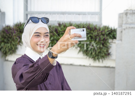 A young Muslim woman in a hijab takes a photo with her smartphone while traveling in the city with her luggage. Travel or vacation concept A young Muslim woman in a hijab takes a photo with her smartphone while traveling in the city with her luggage. Travel or vacation concept 140187291