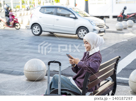 A young Indonesian southeast asian muslim woman sits on a city bench with her suitcase, smiling while using her smartphone as she waits for transport. Travel or vacation concept 140187297