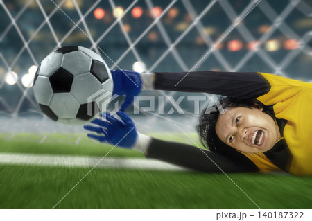 A professional Indonesian southeast asian football or soccer goalkeeper in a yellow jersey dives dramatically to save a ball during an intense football match at the stadium 140187322