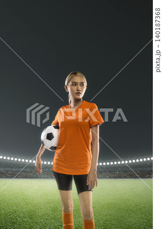 A young Indonesian southeast asian woman soccer or football player in an orange jersey stands on the field of a large stadium at night holding a soccer ball, ready for the game A young Indonesian southeast asian woman soccer or football player in an orange jersey stands on the field of a large stadium at night holding a soccer ball, ready for the game 140187368
