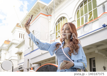 A happy young Indonesian southeast asian woman with auburn hair smiles while taking a selfie with her smartphone outdoors. Travel or vacation concept 140187375