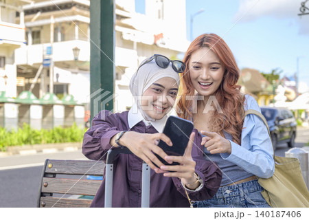 Two young Indonesian southeast asian women, one wearing a hijab, sit on a street bench with a suitcase, smiling while looking at a smartphone together during their travels. Travel or vacation concept Two young Indonesian southeast asian women, one wearing a hijab, sit on a street bench with a suitcase, smiling while looking at a smartphone together during their travels. Travel or vacation concept 140187406