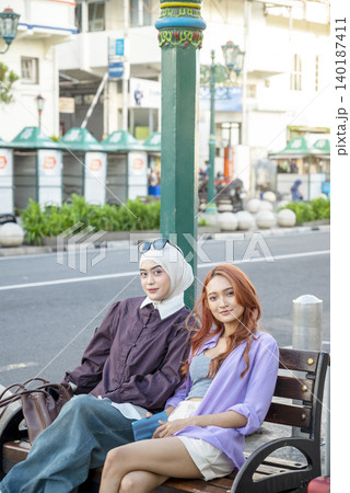 Two stylish young Indonesian southeast asian women, one wearing a hijab and the other with dyed hair, sit on a wooden bench, smiling at the camera. Travel or vacation concept Two stylish young Indonesian southeast asian women, one wearing a hijab and the other with dyed hair, sit on a wooden bench, smiling at the camera. Travel or vacation concept 140187411