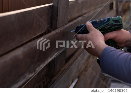man's hands using a green and black cordless electric drill to screw a self-tapping screw into a wooden fence board. The scene captures the practicality of DIY home improvement, woodworking, or constr 140190565