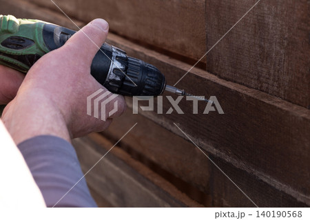 man's hands using a green and black cordless electric drill to screw a self-tapping screw into a wooden fence board. The scene captures the practicality of DIY home improvement, woodworking, or constr man's hands using a green and black cordless electric drill to screw a self-tapping screw into a wooden fence board. The scene captures the practicality of DIY home improvement, woodworking, or constr 140190568