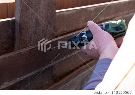 man's hands using a green and black cordless electric drill to screw a self-tapping screw into a wooden fence board. The scene captures the practicality of DIY home improvement, woodworking, or constr 140190569
