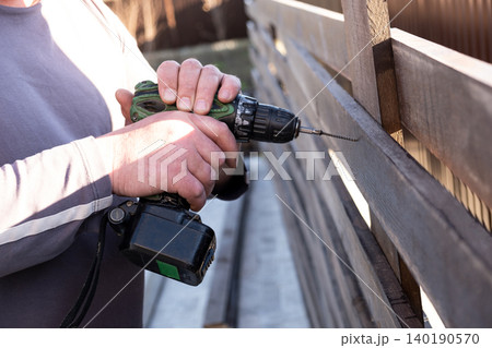man's hands using a green and black cordless electric drill to screw a self-tapping screw into a wooden fence board. The scene captures the practicality of DIY home improvement, woodworking, or constr man's hands using a green and black cordless electric drill to screw a self-tapping screw into a wooden fence board. The scene captures the practicality of DIY home improvement, woodworking, or constr 140190570
