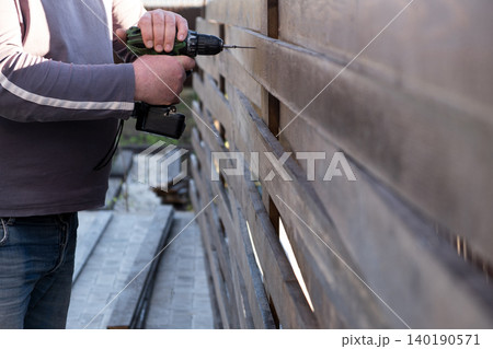 man's hands using a green and black cordless electric drill to screw a self-tapping screw into a wooden fence board. The scene captures the practicality of DIY home improvement, woodworking, or constr 140190571