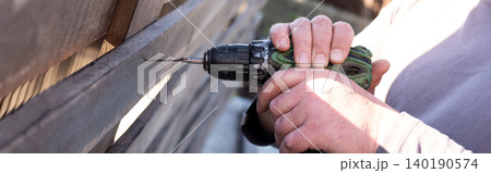 man's hands using a green and black cordless electric drill to screw a self-tapping screw into a wooden fence board. The scene captures the practicality of DIY home improvement, woodworking, or constr 140190574